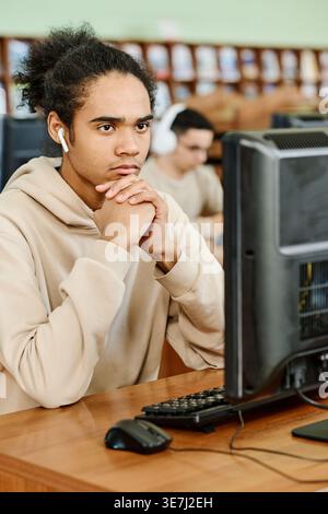 Black male student wearing wireless earphones sitting at desk with clasped hands looking away thinking while studying with computer in university l... Stock Photo