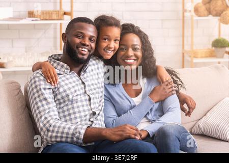 Smiling Black daughter hugging parents from behind sitting together on sofa at home Stock Photo