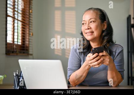 Smiling senior Asian woman holding smartphone looking away while sitting at table with laptop in home office Stock Photo