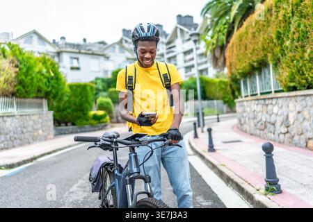 Black man wearing helmet and delivery bag using smartphone delivering food while standing with bicycle outside the house Stock Photo