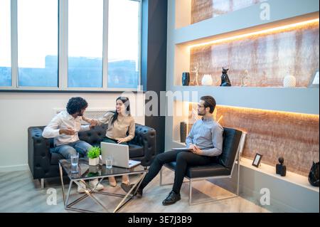 Smiling Asian female and Indian male couple sitting together on sofa near caucasian male psychoanalyst in clinic Stock Photo