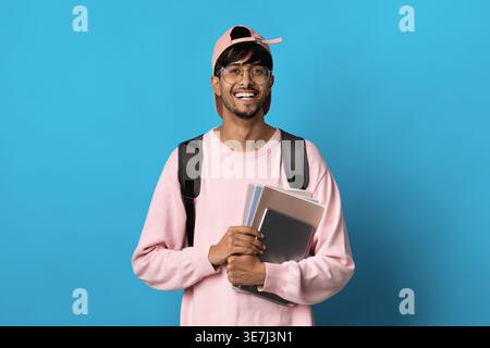 Smiling Middle Eastern male student with backpack and books wearing eyeglasses and pink cap standing against blue background in studio Stock Photo