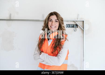 Smiling Hispanic female architect with headphones on neck wearing safety vest with crossed arms standing in construction site during daytime Stock Photo