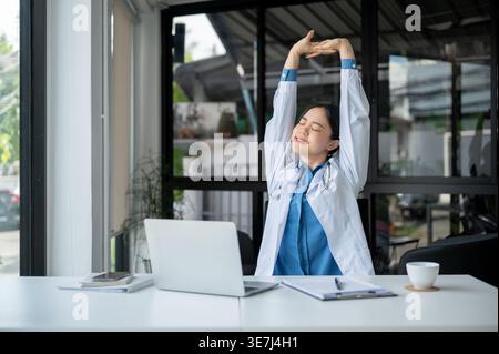 Smiling Asian female doctor with closed eyes raising hands taking break after work sitting at table with laptop in hospital office Stock Photo