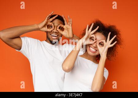 Cheerful Black couple showing okay symbol around eyes while standing against orange background in studio Stock Photo