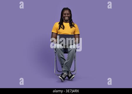Smiling Black man with dreads wearing yellow shirt using laptop sitting on chair against purple background in studio Stock Photo