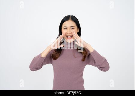 Smiling Asian female with hands near mouth screaming announcement standing against gray background in studio Stock Photo