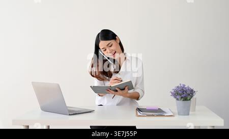 Smiling Asian woman taking notes while having phone call and sitting at table with laptop against white background in studio Stock Photo