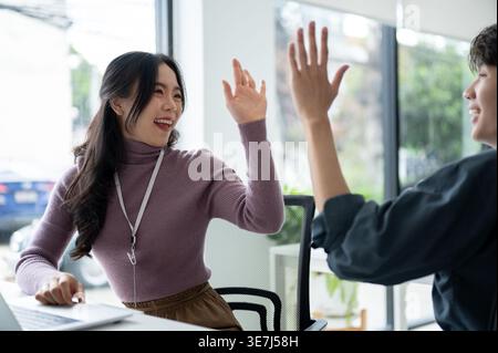 Cheerful Asian female office worker giving high five to male colleague sitting at desk in office Stock Photo