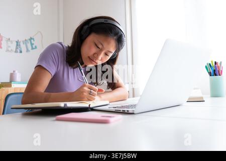 Asian female student in headphones writing on notebook studying while sitting at table with laptop at home Stock Photo