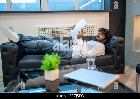 Focused Indian male holding paper reading while lying with crossed legs on sofa in office Stock Photo