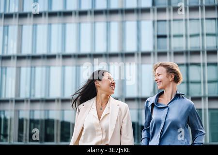 Smiling Asian business woman talking to caucasian female colleague while walking on city street Stock Photo