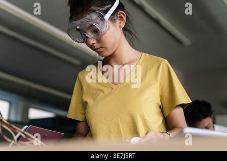Asian female student wearing goggles working on electronics circuit board in classroom Stock Photo
