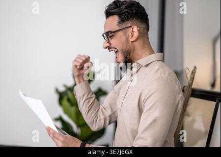 Cheerful Arab man wearing eyeglasses raising arm with closed fist while reading paper standing at home Stock Photo