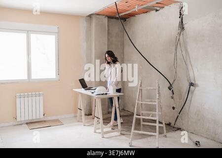 Hispanic female architect having phone conversation standing at table with laptop and hard hat in construction site during daytime Stock Photo