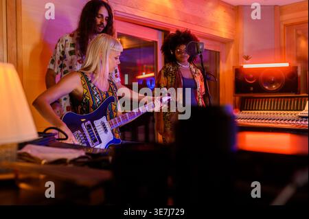 Hispanic female vocalist with afro wearing headphones standing and singing in front of microphone near caucasian male helping female guitarist duri... Stock Photo