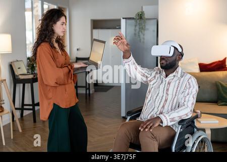 Black bald man with disability wearing virtual reality headset sitting in wheelchair while caucasian female caregiver holding laptop standing in li... Stock Photo