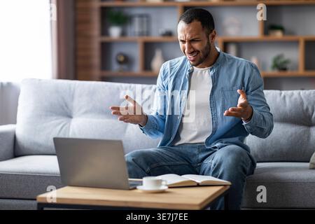 Stressed Black man having problem on laptop while working sitting on sofa in living room at home Stock Photo