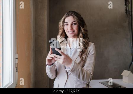 Smiling Hispanic female architect holding smartphone standing near table in construction site during daytime Stock Photo