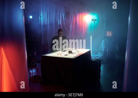 Serious Black man sitting at table with electronic equipment in dark room Stock Photo
