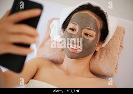 Asian woman with facial clay mask taking selfie lying on bed in spa Stock Photo