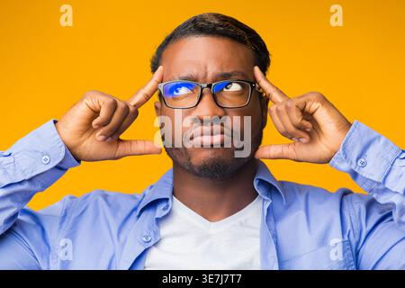 Serious Black man wearing eyeglasses looking away with fingers on sides of his forehead standing against yellow background in studio Stock Photo