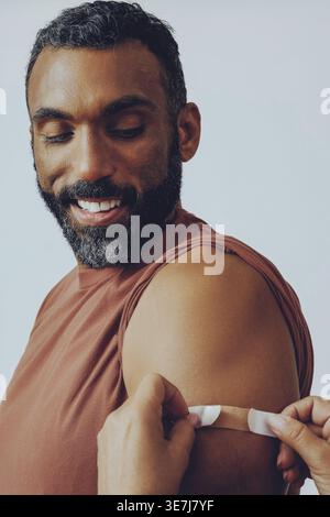 Mixed race bearded man having adhesive bandage placed on his arm standing sideways against white background in studio Stock Photo