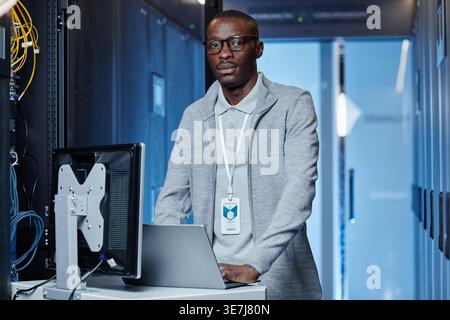 Black male IT engineer with eyeglasses standing at table with laptop in server room in data center Stock Photo