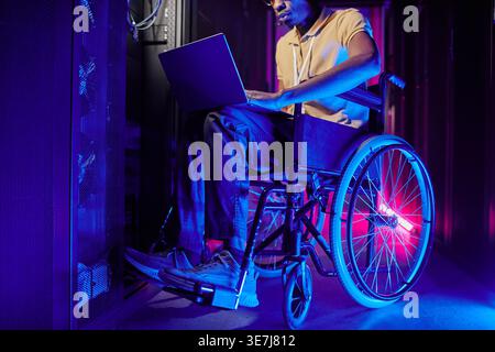 Disabled Black male IT engineer with eyeglasses working on laptop sitting on wheelchair in server room with blue and red neon lights in data center Stock Photo
