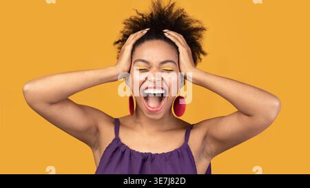 Black woman with closed eyes and open mouth grabbing head standing against yellow background in studio Stock Photo