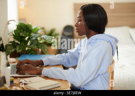 Black female student typing on her laptop sitting at study table beside potted plant near bed inside her dorm room during daytime Stock Photo