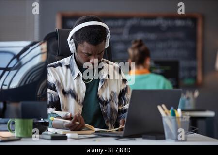 Black male programmer wearing headphones holding pen and reading his planner while sitting at table with laptop near caucasian female colleague in ... Stock Photo