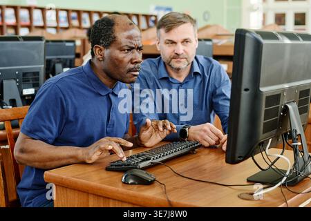 Middle-aged Black male immigrant sitting at desk with caucasian male instructor helping him to understand task on computer in library Stock Photo