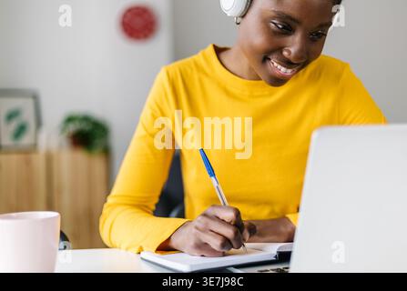 Smiling Black female student in headphones sitting at table with laptop writing notes during online class at home Stock Photo