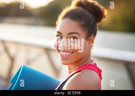 Smiling Mixed race woman holding yoga mat standing on seaside promenade during sunset Stock Photo