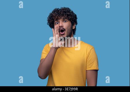 Indian man with curly hair standing with hand near mouth calling someone against blue background Stock Photo