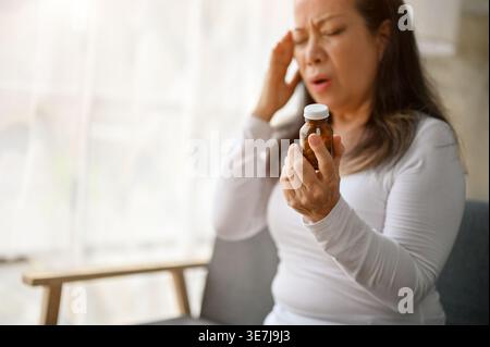 Senior Asian woman touching temple holding medicine bottle having headache sitting on sofa at home Stock Photo