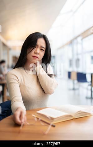 Tired Asian woman suffering from neck pain sitting at table with book in cafe Stock Photo