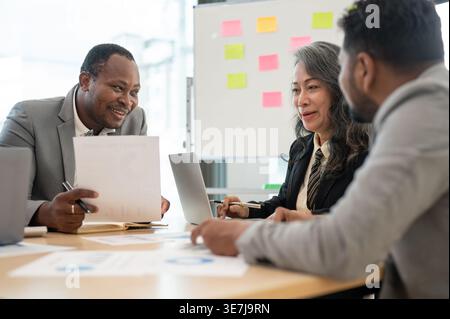 Smiling senior Asian business woman discussing project details to Black and Indian male colleagues while sitting at table in the office Stock Photo