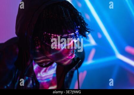 Serious Black man with afro wearing hoodie and led augmented reality glasses looking away while standing in dark room with colorful neon lights Stock Photo