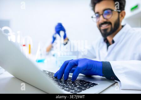 Cheerful Indian male scientist wearing lab coat and latex gloves holding glass dropper with reagents and making notes on laptop while sitting at hi... Stock Photo