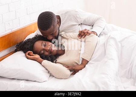 Black man waking up his wife with kiss on cheek lying on bed in bedroom at home Stock Photo