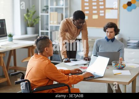 Black male IT specialist talking to male and female colleagues working on laptop sitting at table in office Stock Photo
