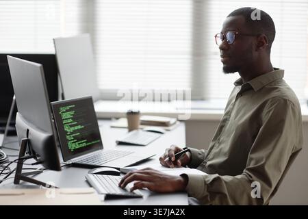 Side view of Black man in glasses holding pen typing on keyboard running data analysis on computer and laptop at desk sitting in office Stock Photo