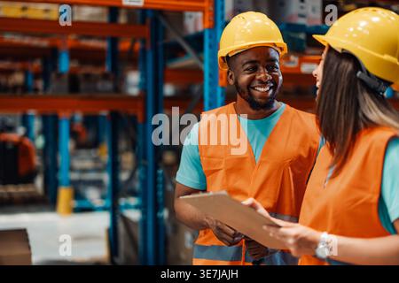 Black male worker wearing hard hat and safety vest checking inventory list with caucasian female coworker holding clipboard while standing in wareh... Stock Photo