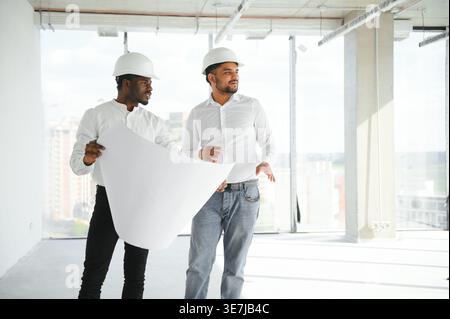 Diverse group of male engineers wearing hard hats standing together and looking at blueprint during work on construction site Stock Photo
