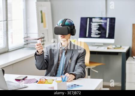 Asian male programmer wearing virtual reality headset while sitting at table with eyeglasses, laptop, and smartphone in office Stock Photo
