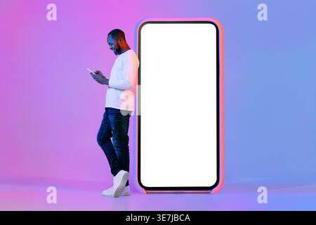 Smiling Black man using smartphone leaning on huge phone while standing against purple background in studio with neon lights Stock Photo