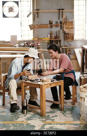 Asian male and female carpenters sharing smLifestylephones while having coffee break sitting on stool in workshop during daytime Stock Photo