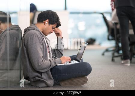 Stressed mixed-race male employee wearing earphones touching face using laptop leaning against glass wall sitting with closed eyes and crossed legs... Stock Photo